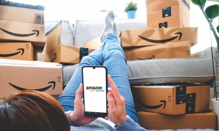 Una persona tumbada en el suelo apoya los pies sobre una pila de paquetes de Amazon Prime mientras sostiene un teléfono móvil en el que está abierta la aplicación Amazon Prime
